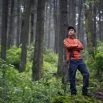 A hiker stands confidently in a lush forest, arms crossed, portraying exploration.