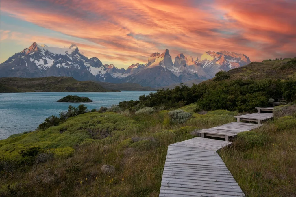 Wooden boardwalk leading to Torres del Paine mountains under a vibrant sunset sky in Chile.